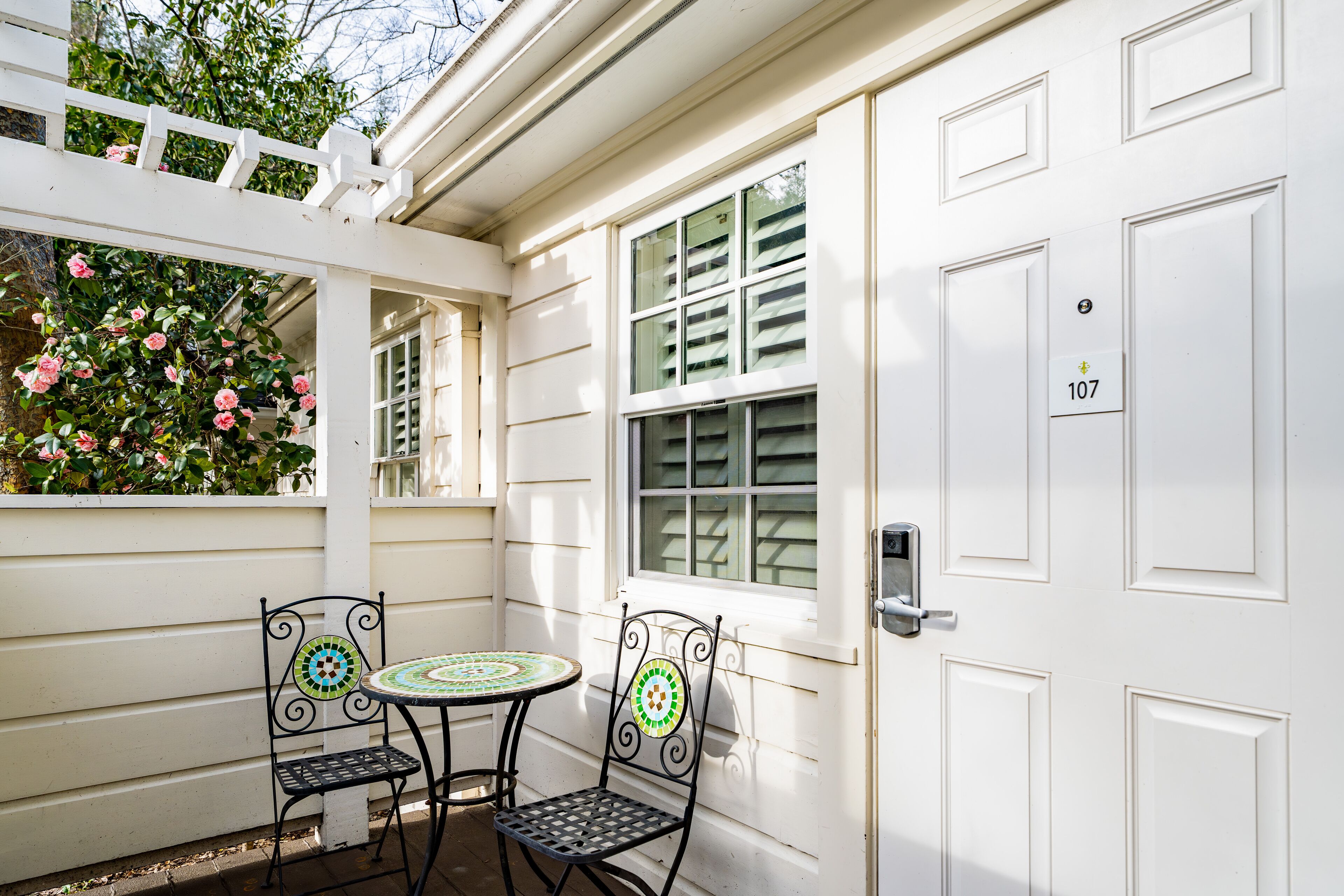 Una puerta blanca con el número 107, una ventana con contraventanas y un pequeño patio con dos sillas y una mesa.