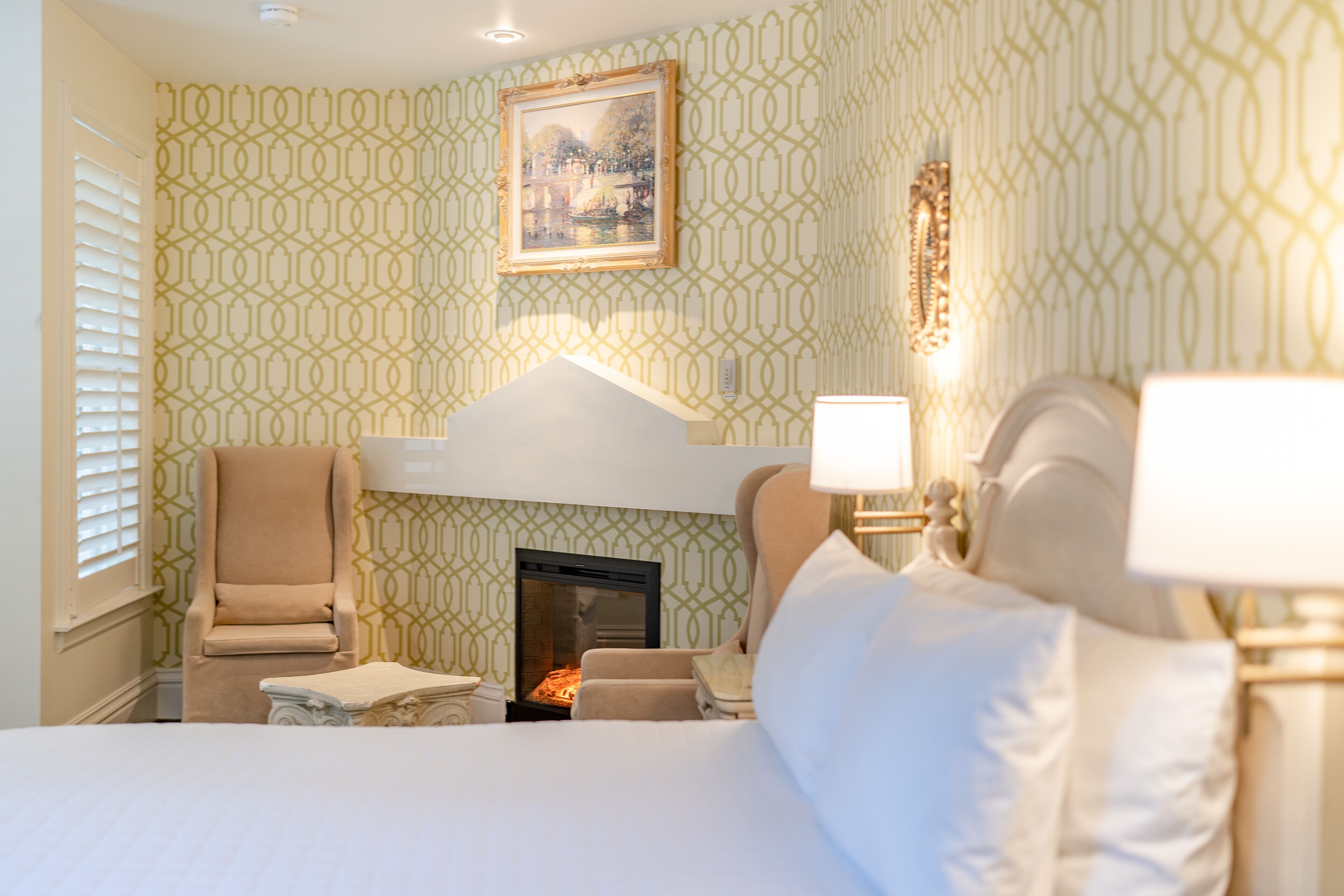 A hotel room with a fireplace, a bed, two armchairs, and a painting on the wall.