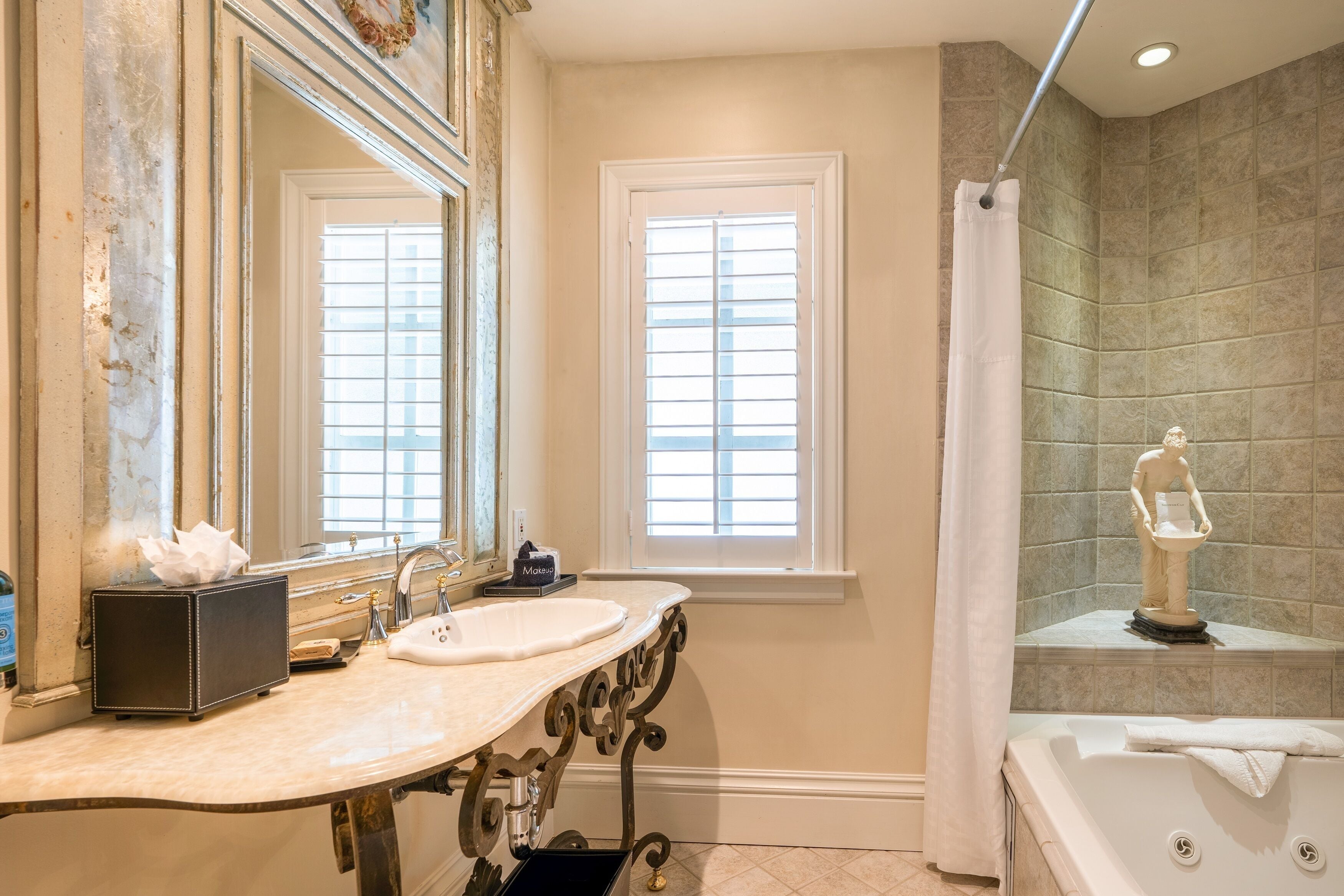 A bathroom with a marble sink, two windows with blinds, a shower with a statue, and a bathtub.