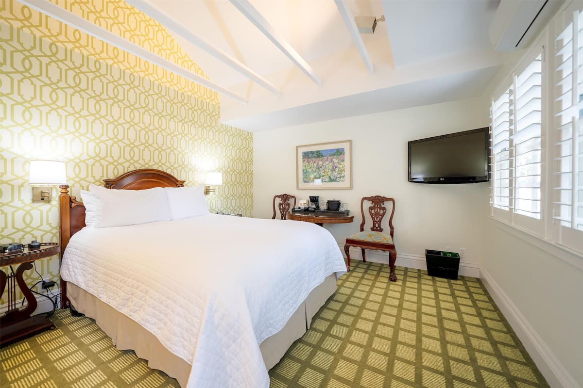 A hotel room with a large bed, a bedside table with a lamp, a small table with a chair, a painting on the wall, and a TV mounted on the wall.