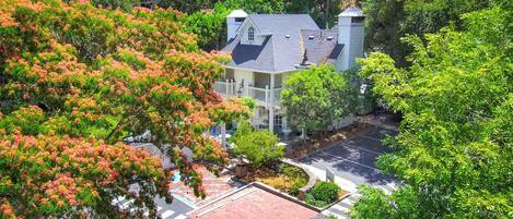 A large, two-story house with a prominent balcony, surrounded by lush greenery and a paved driveway.