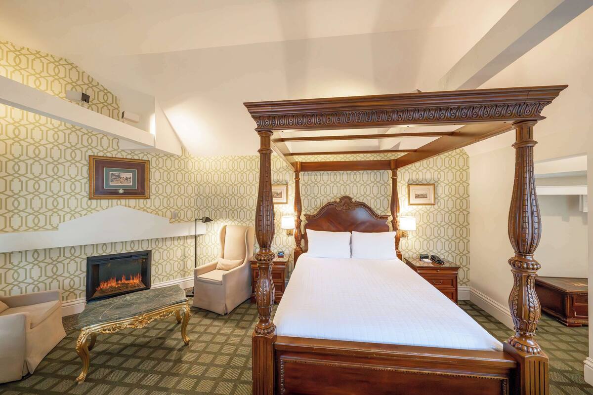 A four-poster bed with a wooden frame, a fireplace, a chair, and a nightstand with a lamp.