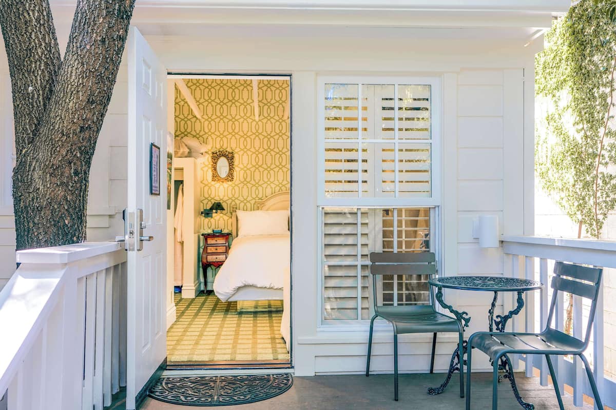 A room with a bed, a small table, and a chair. There is a window with blinds and a tree outside.