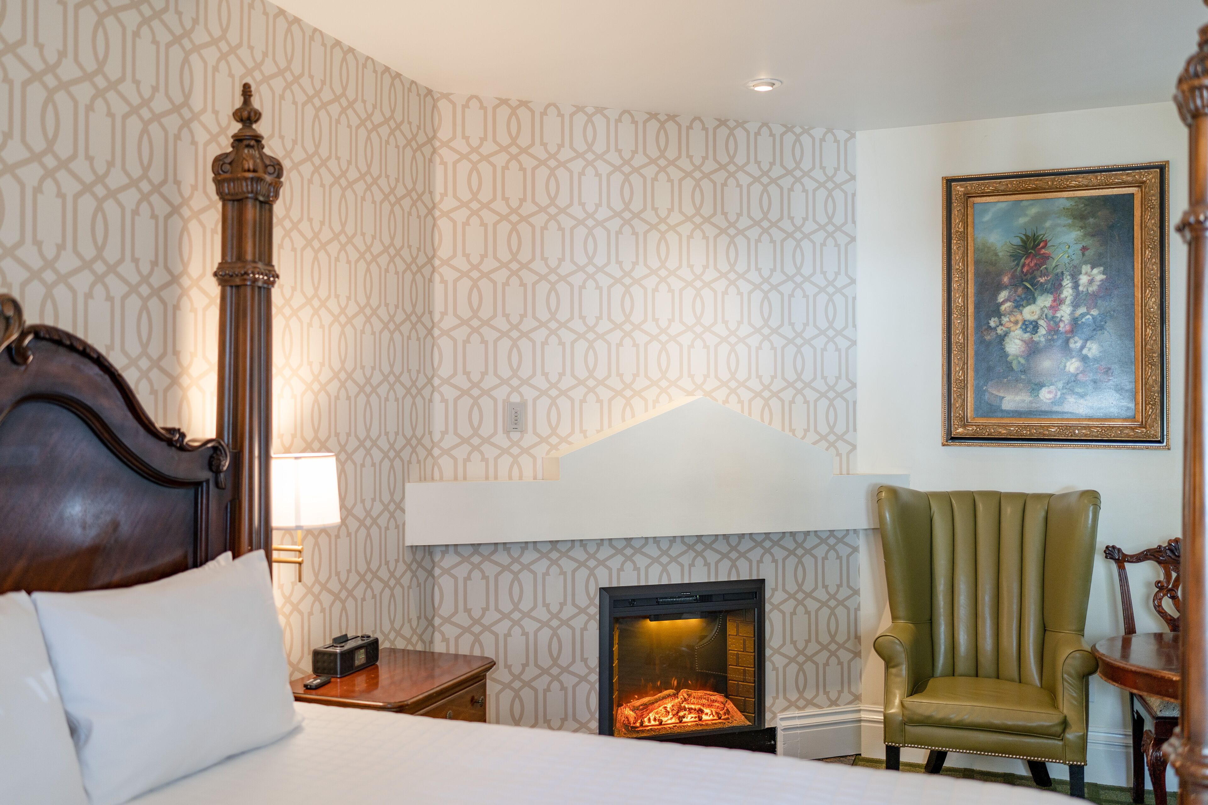 Una habitación de hotel con cama, mesita de noche, sillón verde, chimenea y un cuadro en la pared.