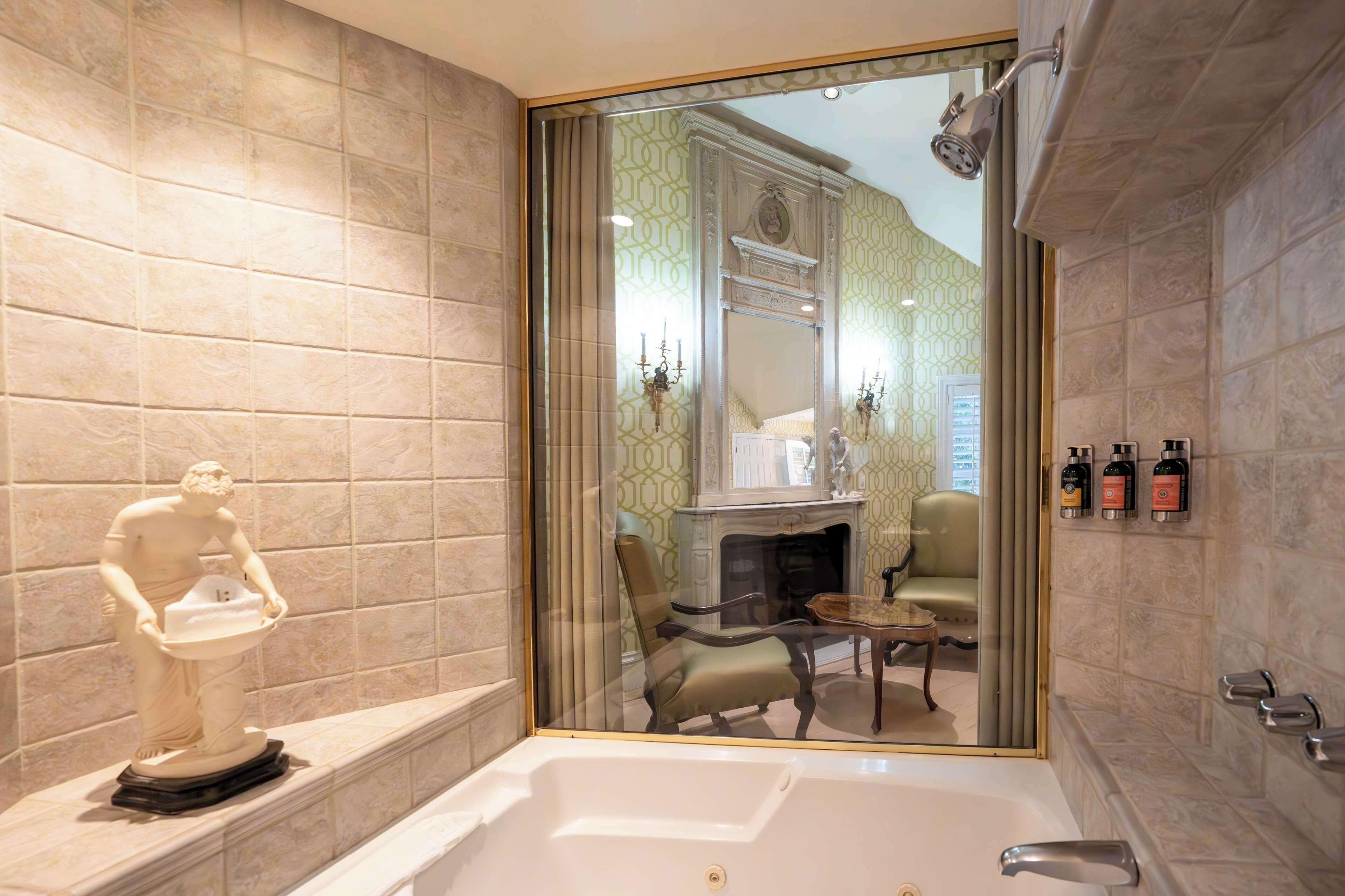 A bathroom with a bathtub, a marble statue, a mirror, a fireplace, and toiletries on the wall.