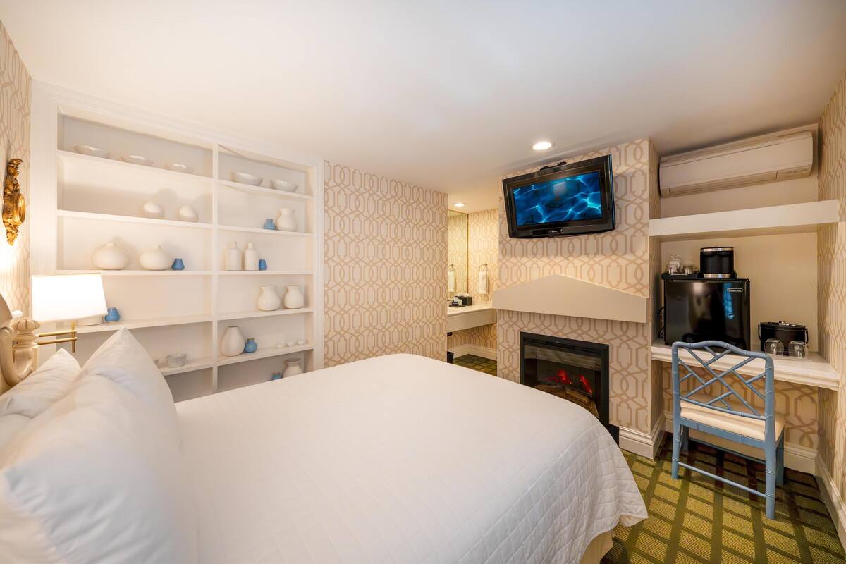 A hotel room with a large bed, a television mounted on the wall, a fireplace, and a built-in shelf with various items.