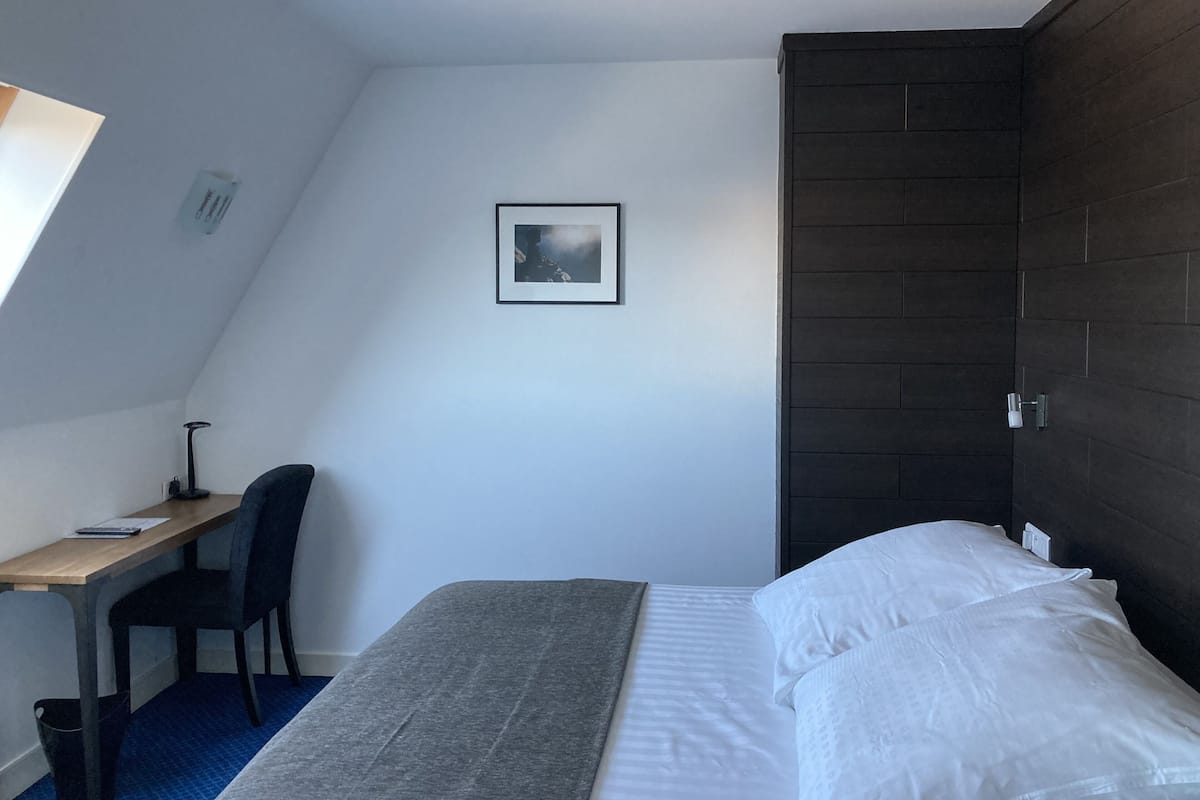 Ein kleines, modernes Hotelzimmer mit Bett, Schreibtisch, Stuhl und einer dunklen Holzwand.