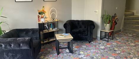 A living room with two black armchairs, a small table, a bookshelf, and a floral-patterned carpet.