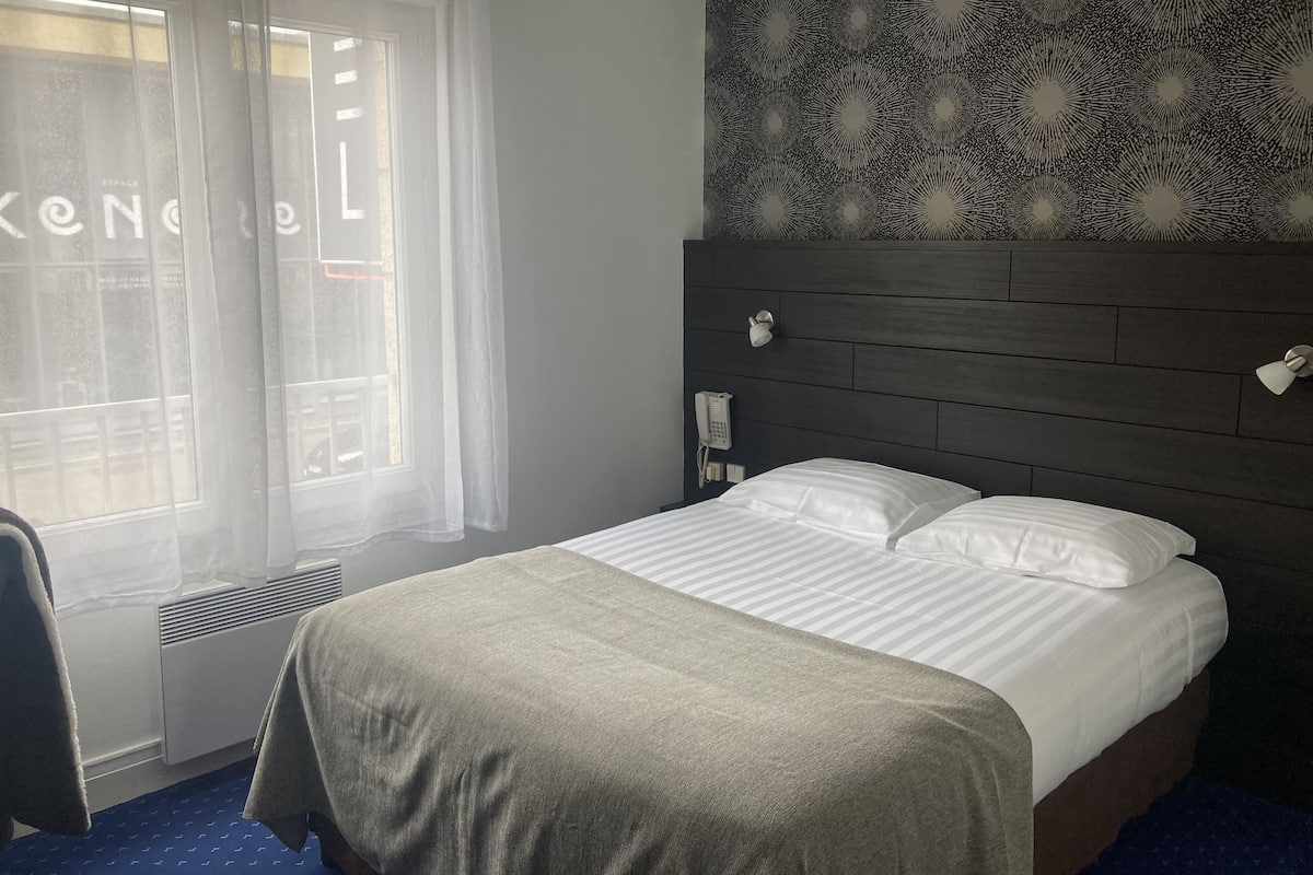 Ein Hotelzimmer mit Bett, Nachttischtelefon, Fenster mit durchsichtigen Vorhängen und einer Wand mit Muster.