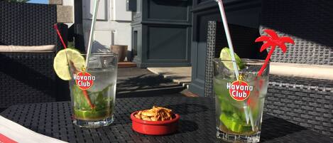 Two drinks with straws and lime slices on a table, a red bowl of nuts, and a newspaper.