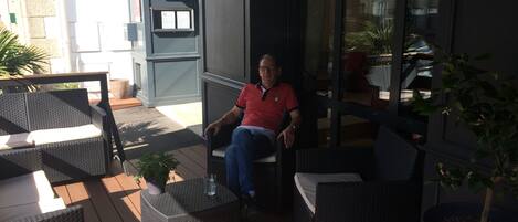 Un hombre sentado en una silla de mimbre en un patio al aire libre con mesas y sillas de mimbre, una pequeña planta en maceta y un vaso de agua sobre una de las mesas.