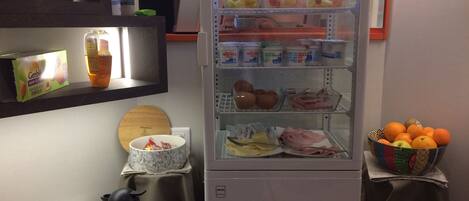 A kitchen area with a refrigerator stocked with fruits, a coffee maker, and a bowl of oranges.