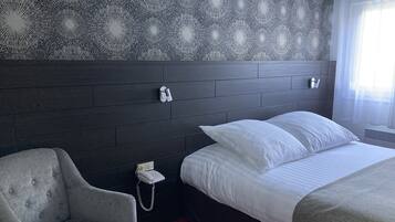 Habitación de hotel con tablero de cabecera de madera oscura, una cama individual con ropa de cama blanca, un sillón gris y un teléfono en la mesita de noche.