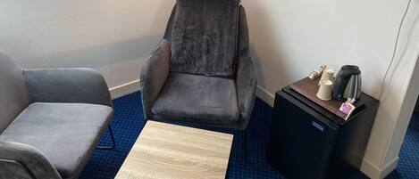 Una habitación con un sillón gris, una mesa de centro de madera y un pequeño armario negro con una tetera y una taza.