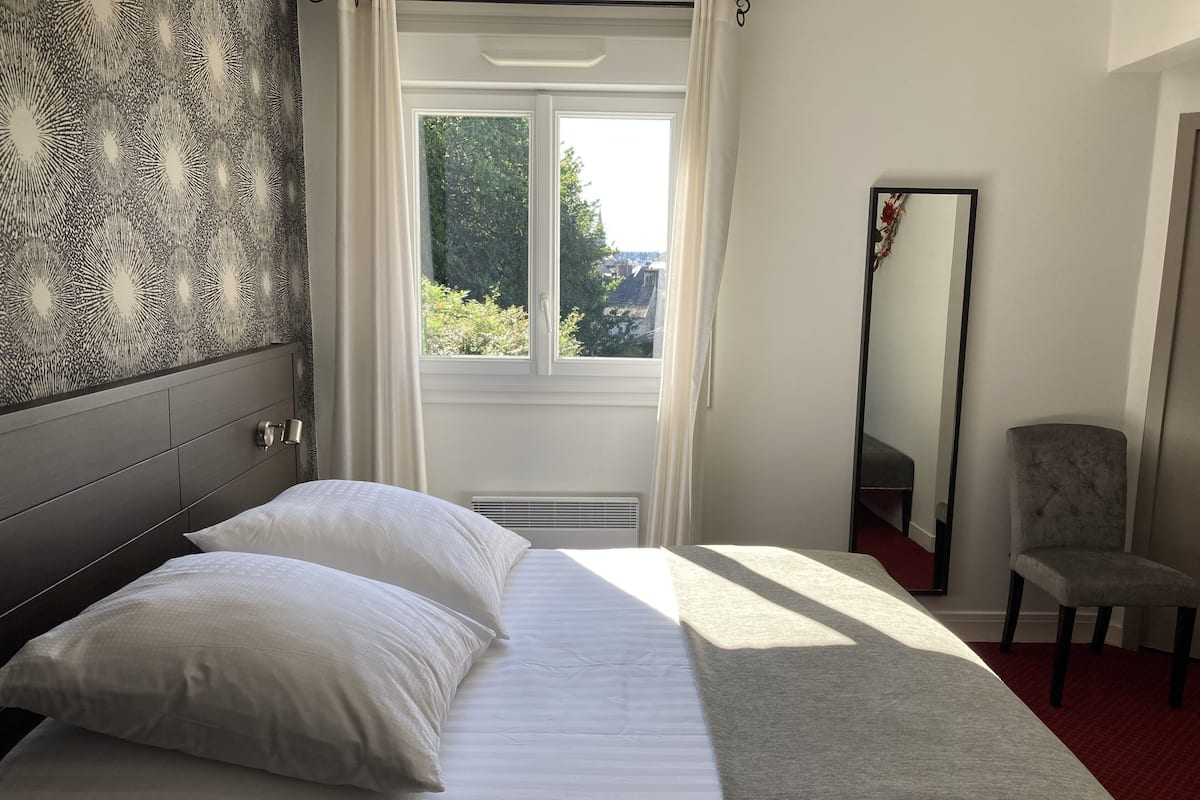 Ein Schlafzimmer mit Bett, Spiegel und Blick auf Grünfläche.