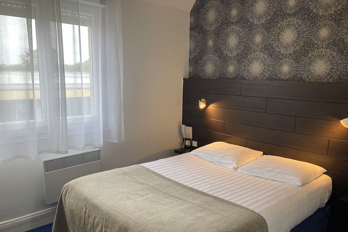 Ein Hotelzimmer mit einem Bett, einem Kopfteil mit Wandleuchten, einem Telefon und einem Fenster mit durchsichtigen Vorhängen.