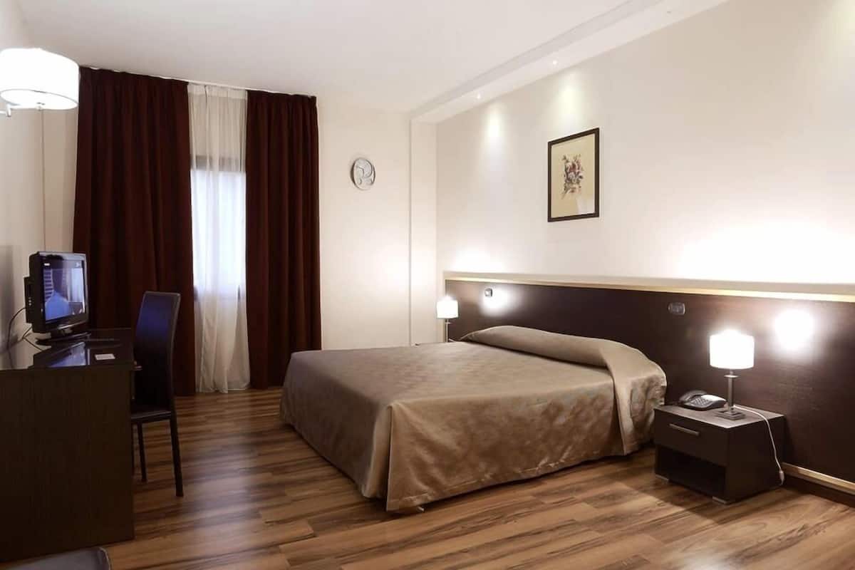 A hotel room with a bed, bedside table, lamp, desk with a computer, and a chair.