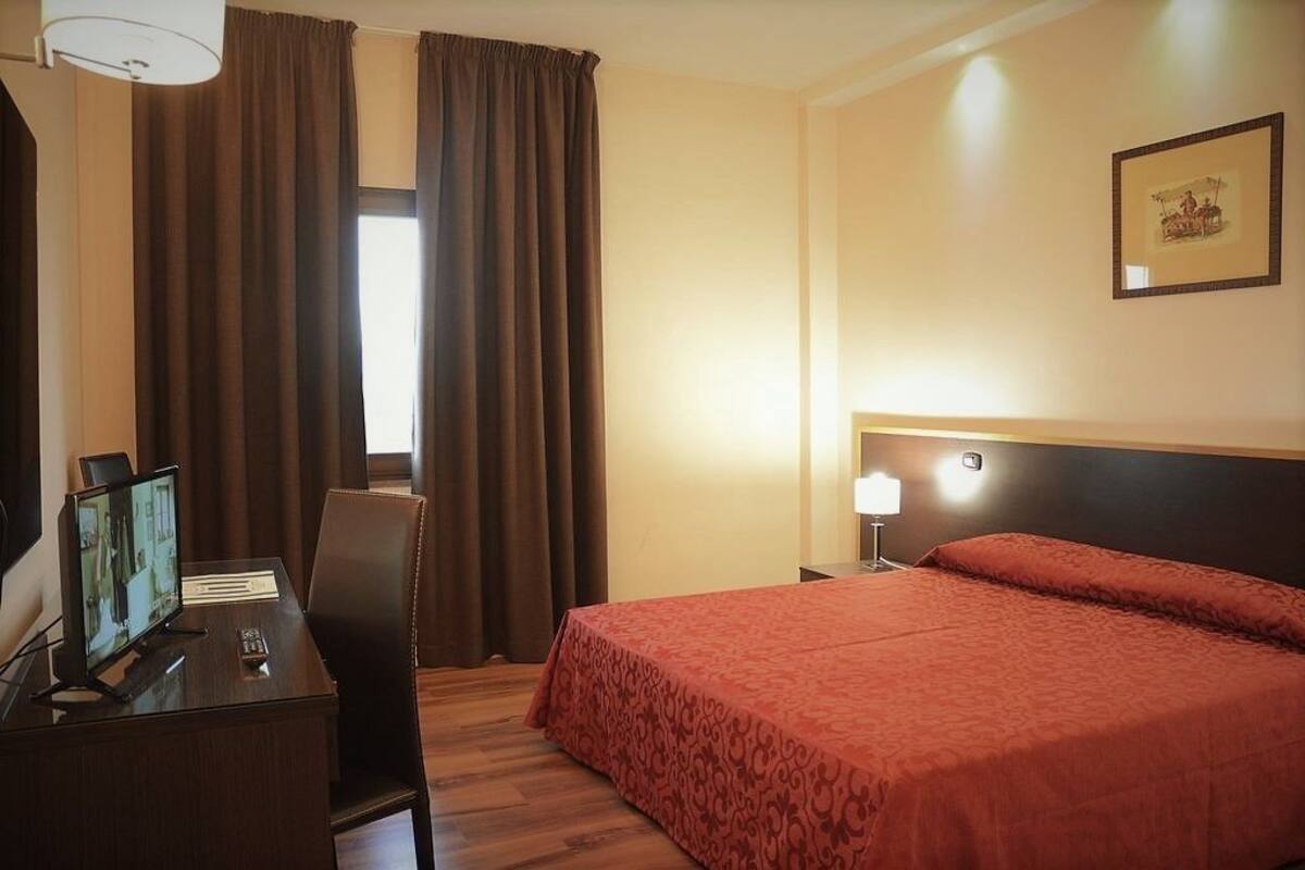 A hotel room with a bed, a desk with a computer, a chair, a window with curtains, and a wall-mounted lamp.