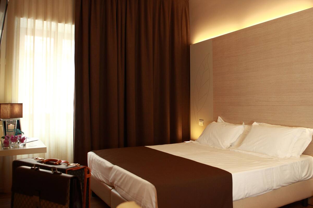 Een hotelkamer met een groot bed, een bureau met een lamp en een vaas met bloemen op het nachtkastje.