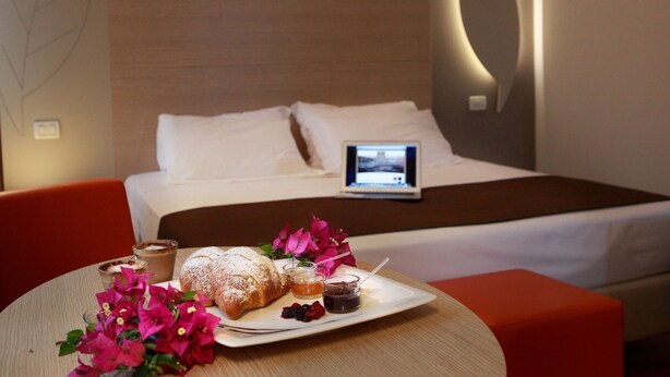 Een hotelkamer met een bed, een nachtkastje met een laptop, een ontbijttafel met gebak en jam, en een bloevas.