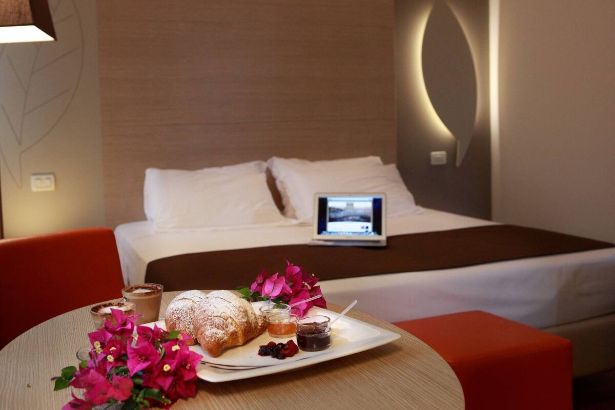 Een hotelkamer met een bed, een nachtkastje met een laptop, een ontbijttafel met gebak en jam, en een bloevas.