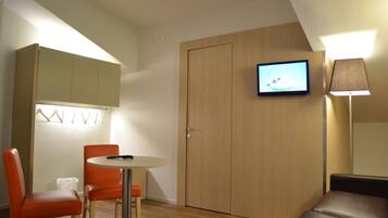 A small, modern room with a wooden door, a television mounted on the wall, a round table with two orange chairs, and a floor lamp.