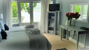 A hotel room with two beds, a TV on a stand, a desk with a vase of flowers, and a view of greenery outside. - Roseroc Boutique Guest House (Knysna)