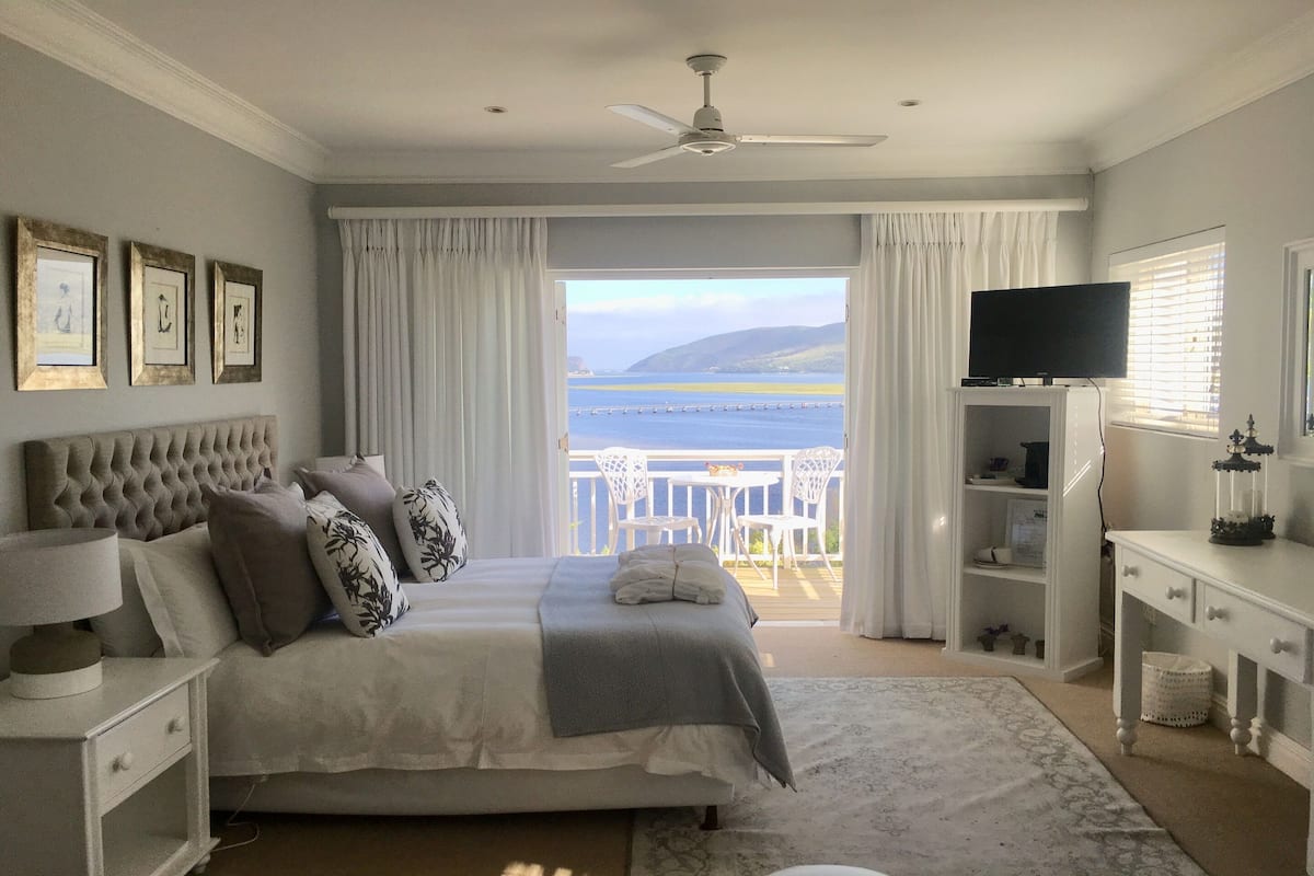 A bedroom with a bed, a nightstand, a TV, a dresser, and a view of the sea.