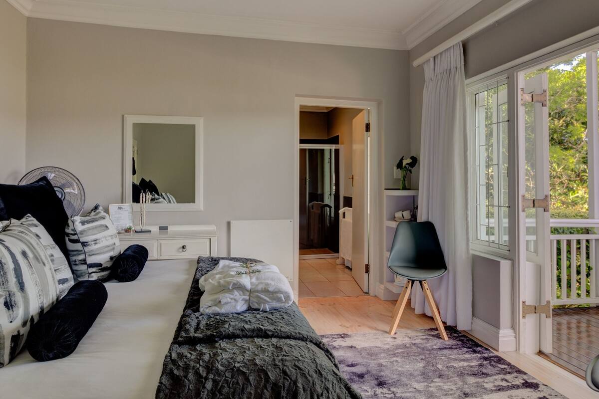 A bedroom with a bed, bedside table, and a chair. There is a door leading to a bathroom and a balcony with a railing.
