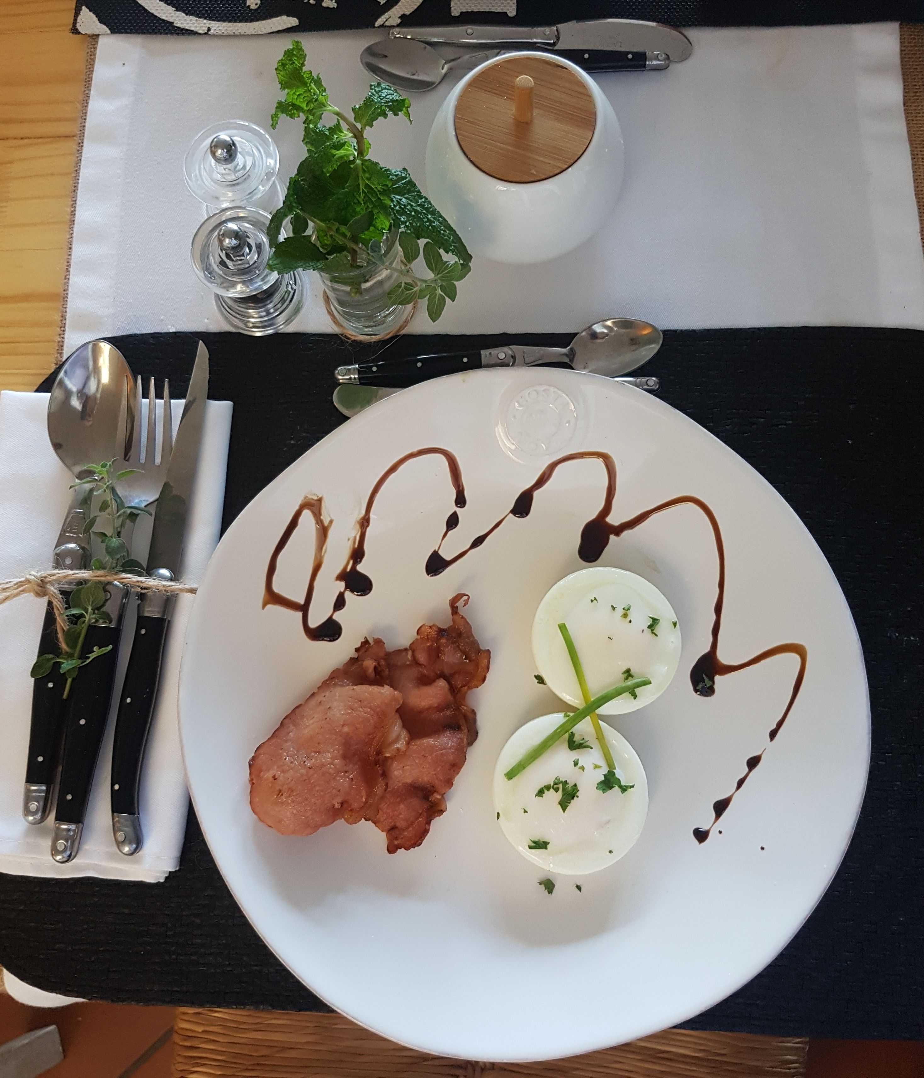 A plate of food with bacon, boiled eggs, and a sauce, accompanied by cutlery and a cup on a table.