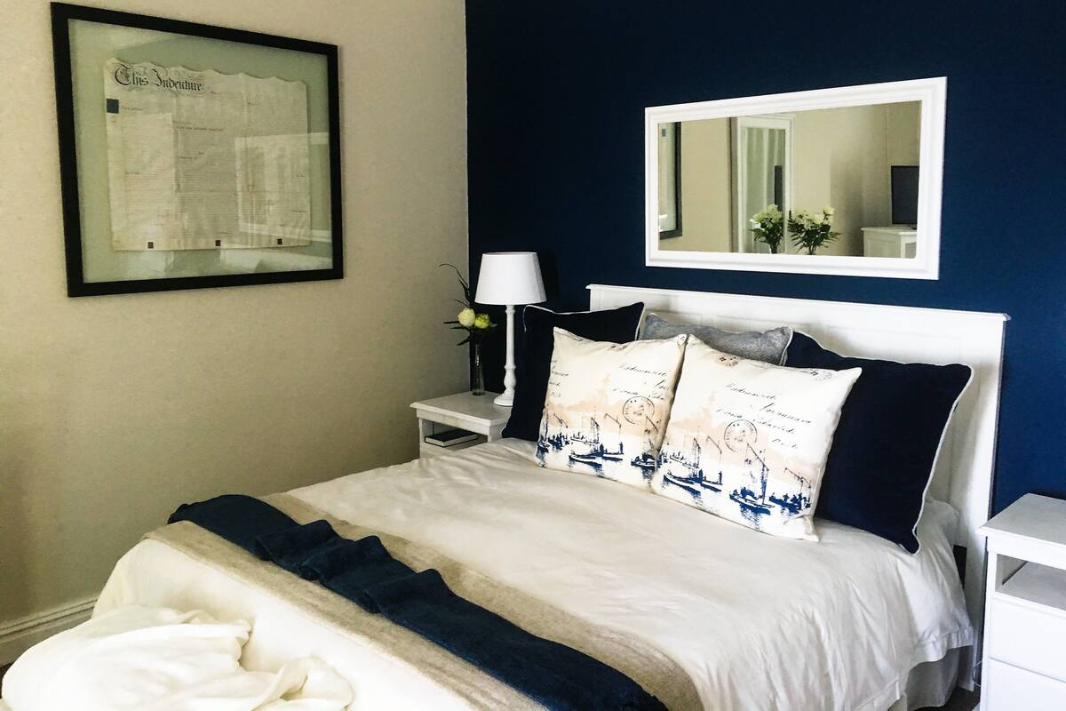 A neatly made bed with a white and navy blue bedspread, a framed document on the wall, a nightstand with a lamp, and a mirror above the bed.