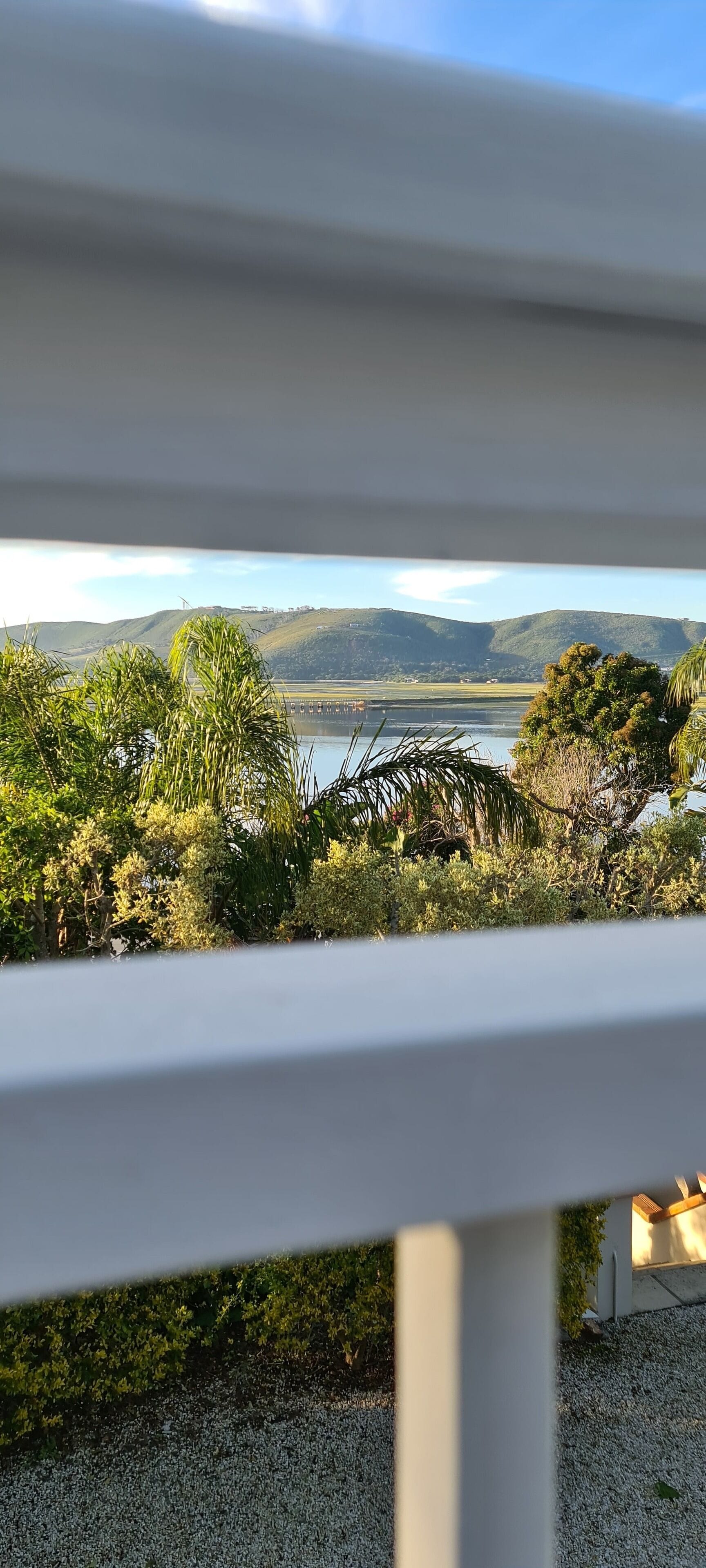 A view from a balcony with a railing, overlooking a body of water, hills, and a pergola structure.
