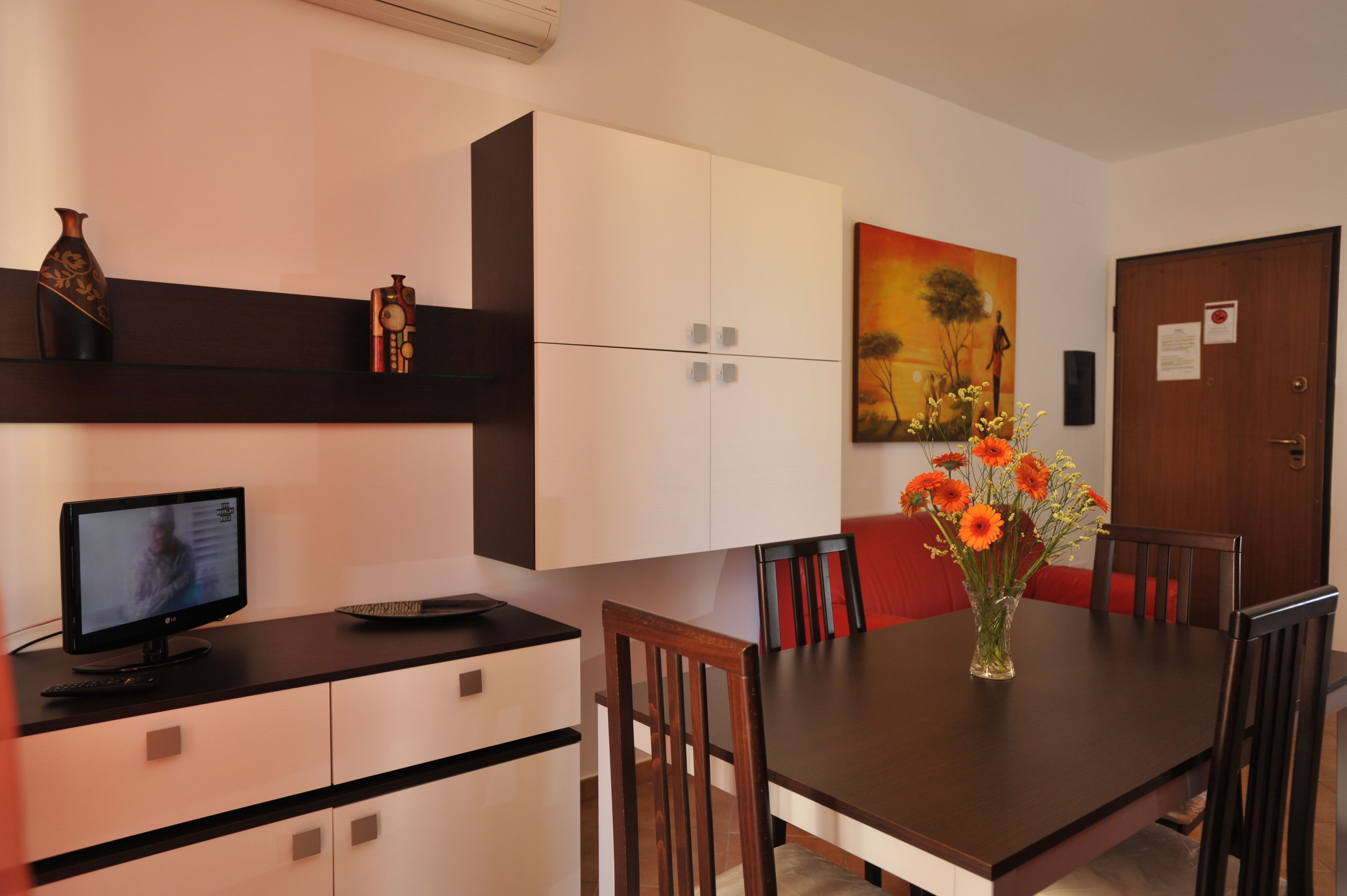 A modern dining area with a dark wooden table, red chairs, and a vase of orange flowers. There is a television on a black stand to the left.