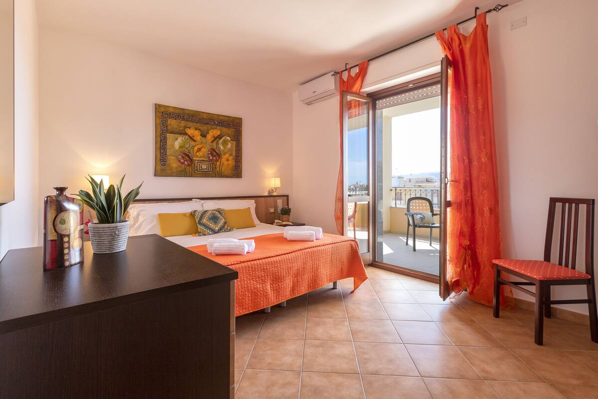 A hotel room with a bed, a desk, a chair, a potted plant, and a balcony with orange curtains.
