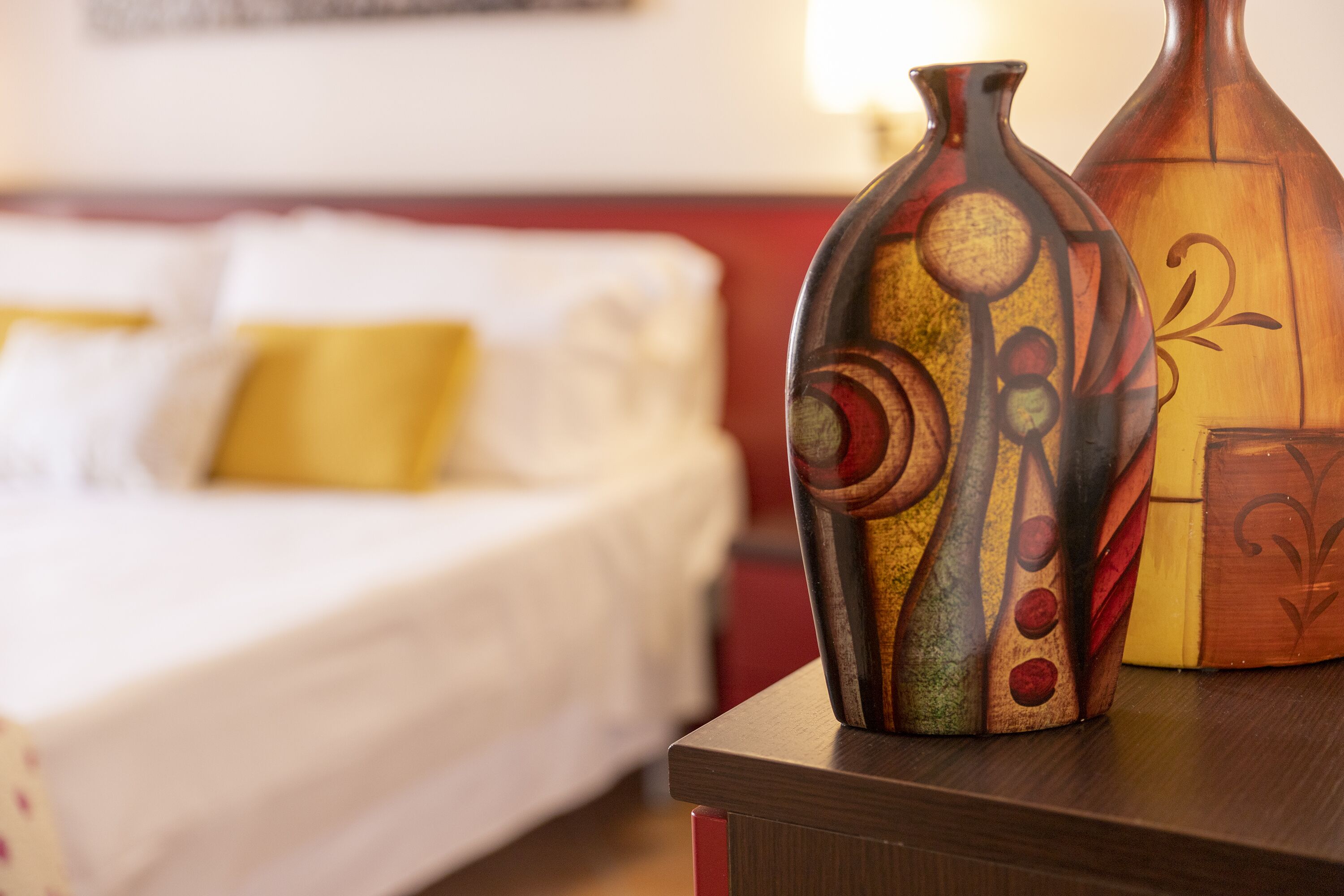 Cama com roupa de cama branca e um travesseiro amarelo, mesa de cabeceira de madeira com dois vasos decorativos, e uma parede com um quadro emoldurado.