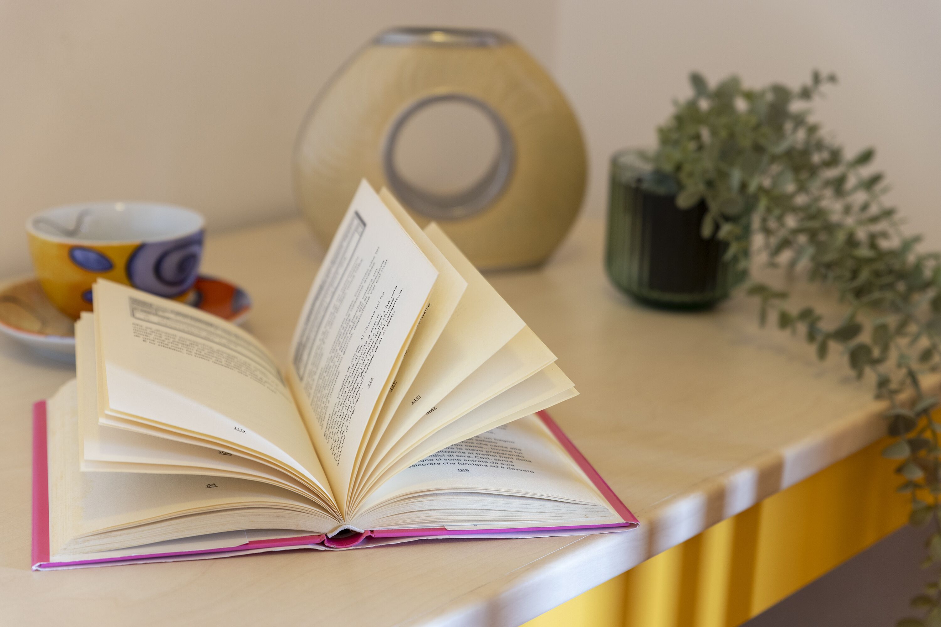 An open book on a yellow table with a pink bookmark, a yellow cup with a purple design, a wooden vase, and a green plant.