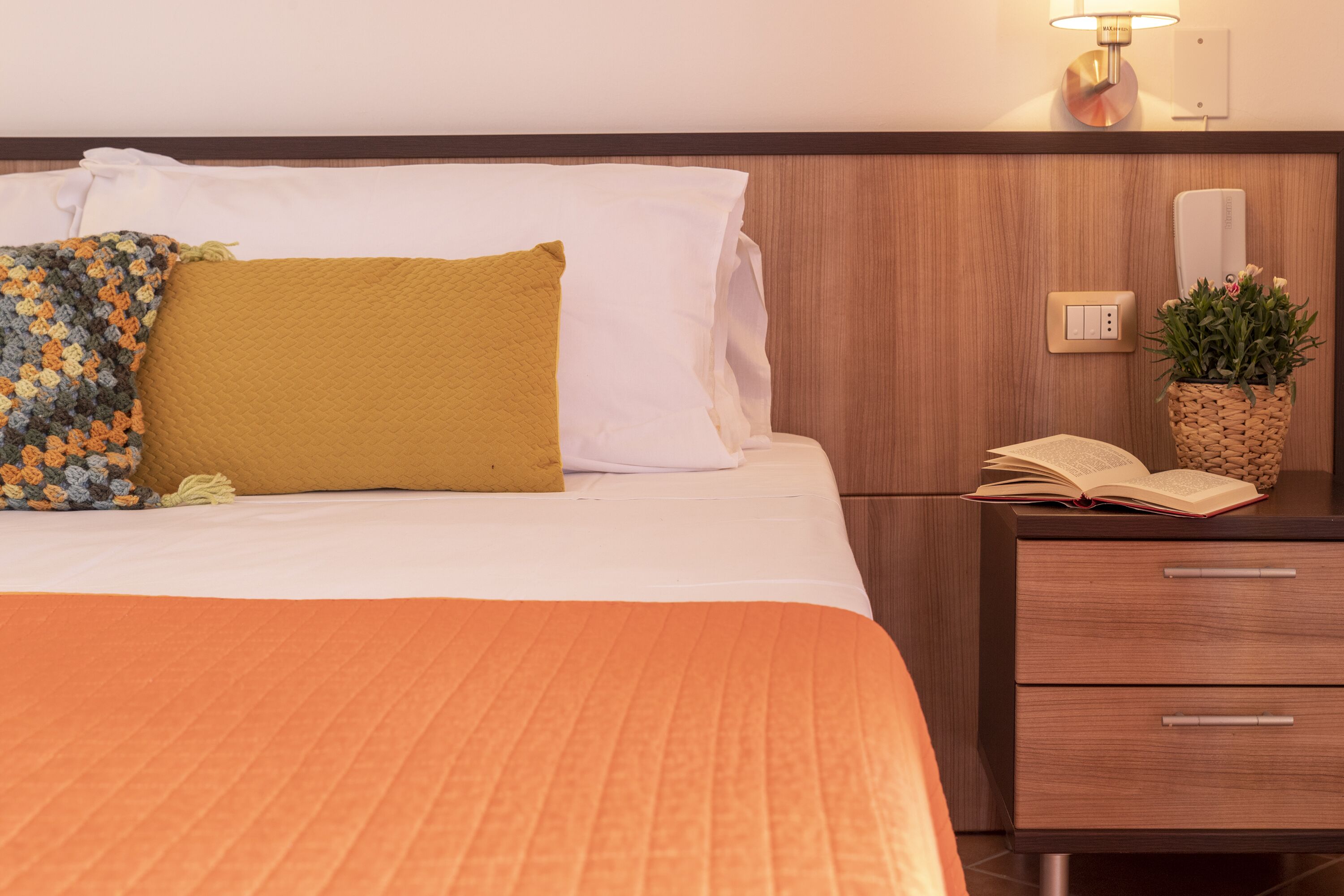 A neatly made bed with a white and mustard-colored pillow, an orange bedspread, and a small bedside table with a book and a potted plant.