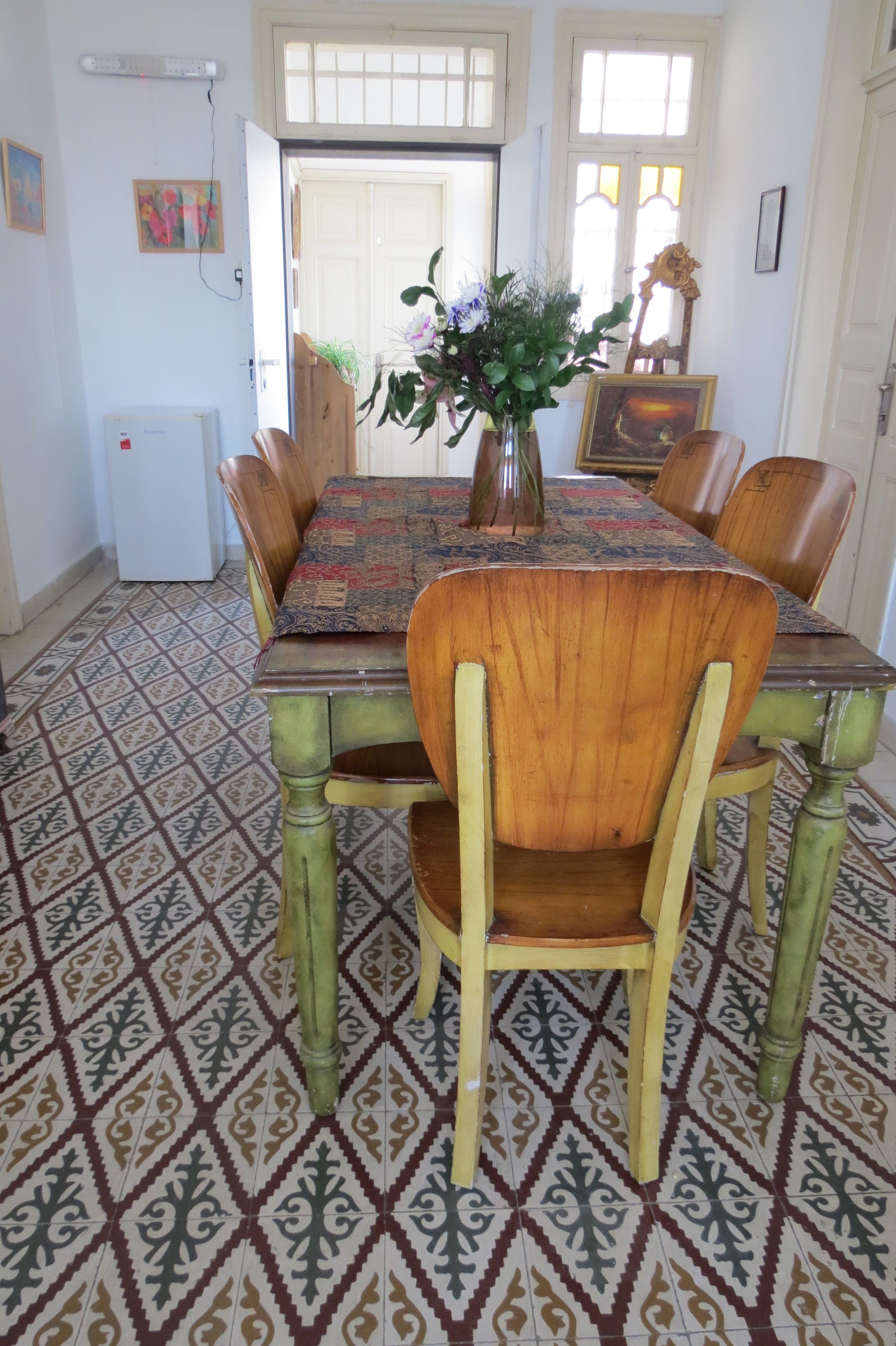 Et spiseområde med et mønstret gulv, et bord med dug i et mønster, træstole, en vase med blomster, og et køleskab i baggrunden.