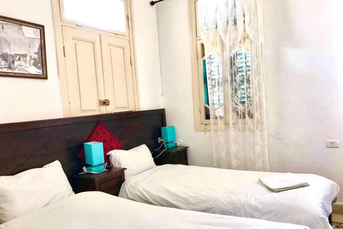 A hotel room with two beds, a wooden headboard, a window with lace curtains, a small table with a blue lamp, and a framed picture on the wall.