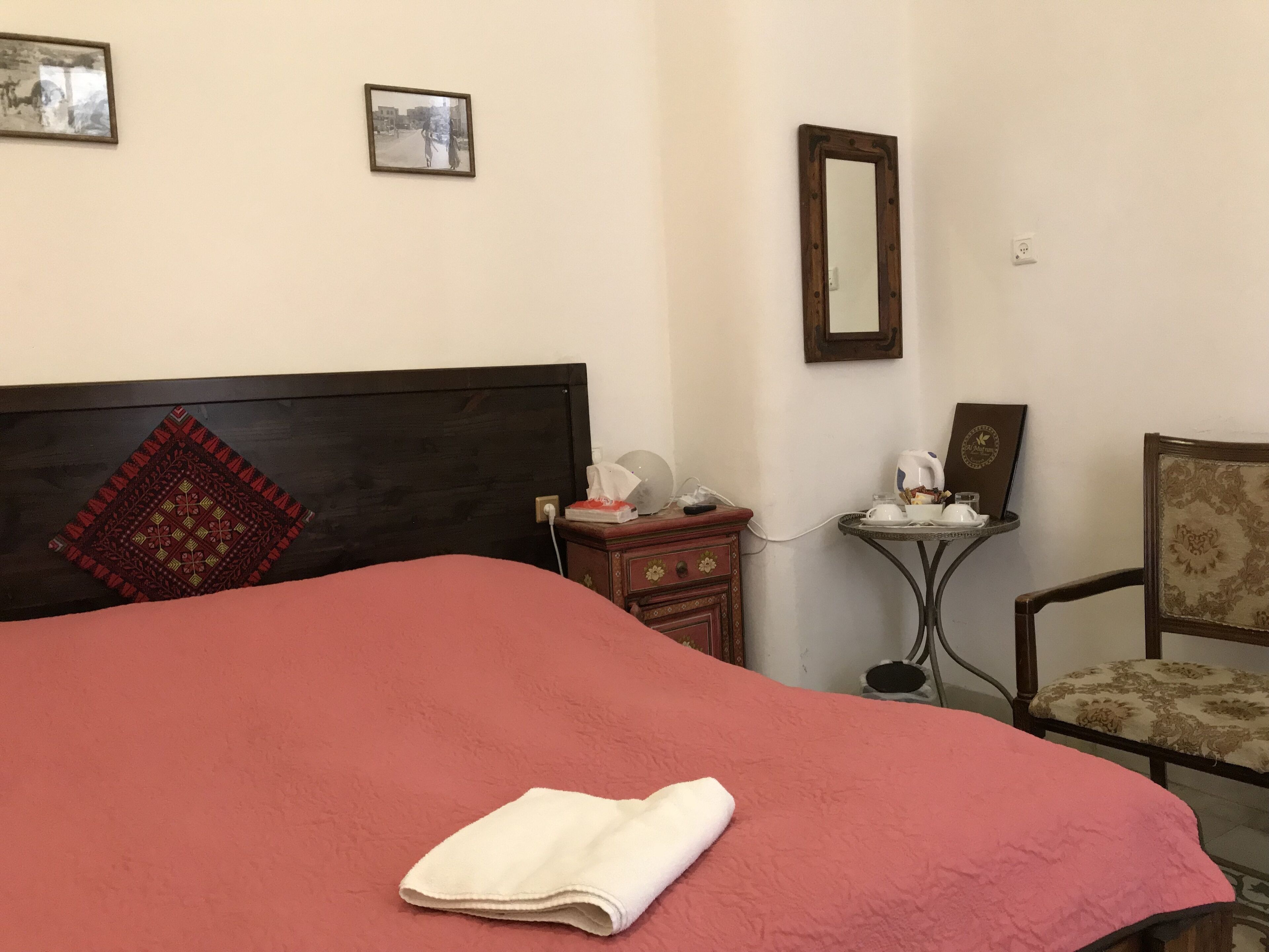 Una cama bien hecha con una manta roja y una toalla blanca, un cabecero de madera, una mesita de noche con un pañel de papel y una lámpara, una silla con un cojín estampado, y dos cuadros en la pared.