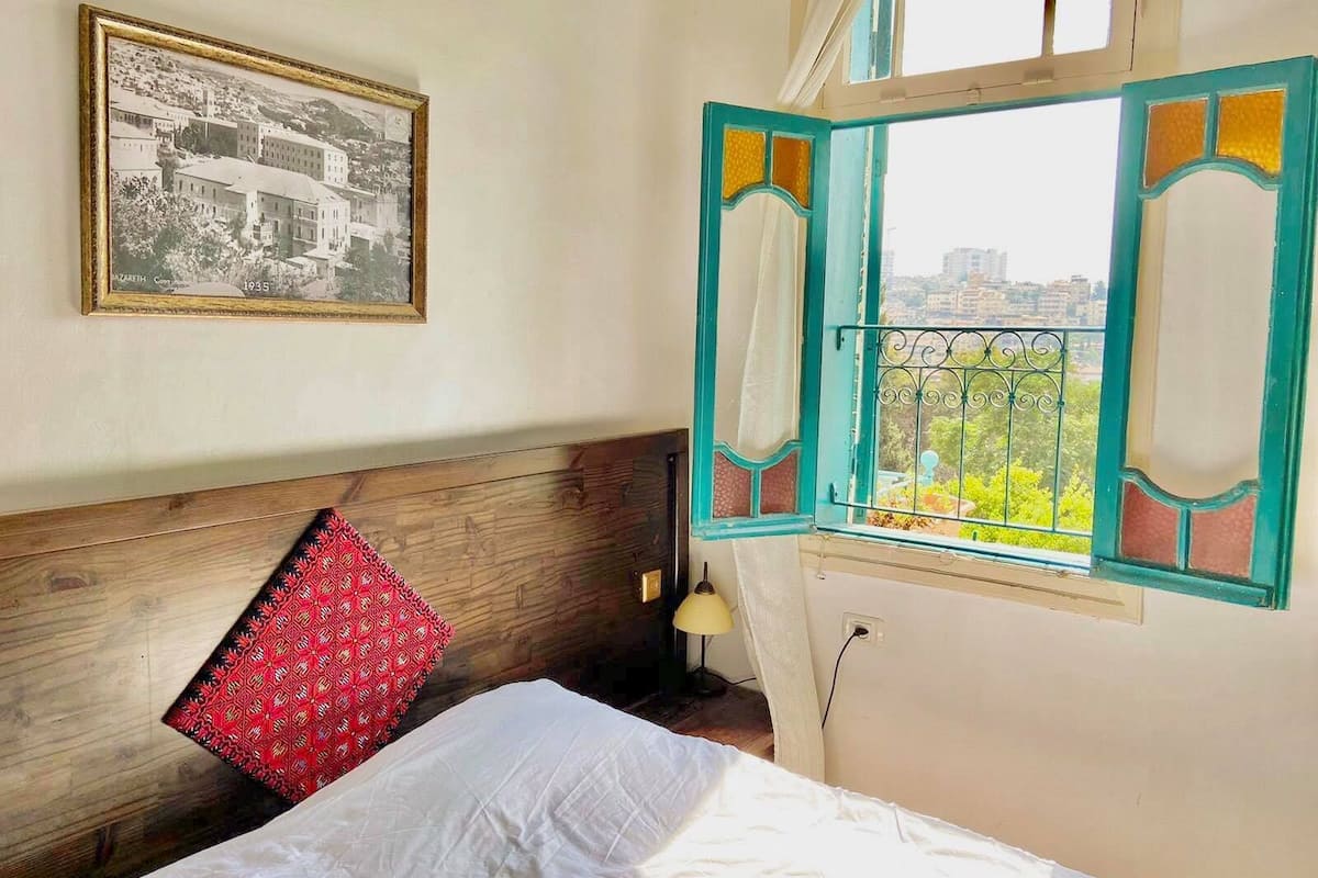 A bedroom with a bed, a window with a view, a wall-mounted picture, and a bedside lamp.