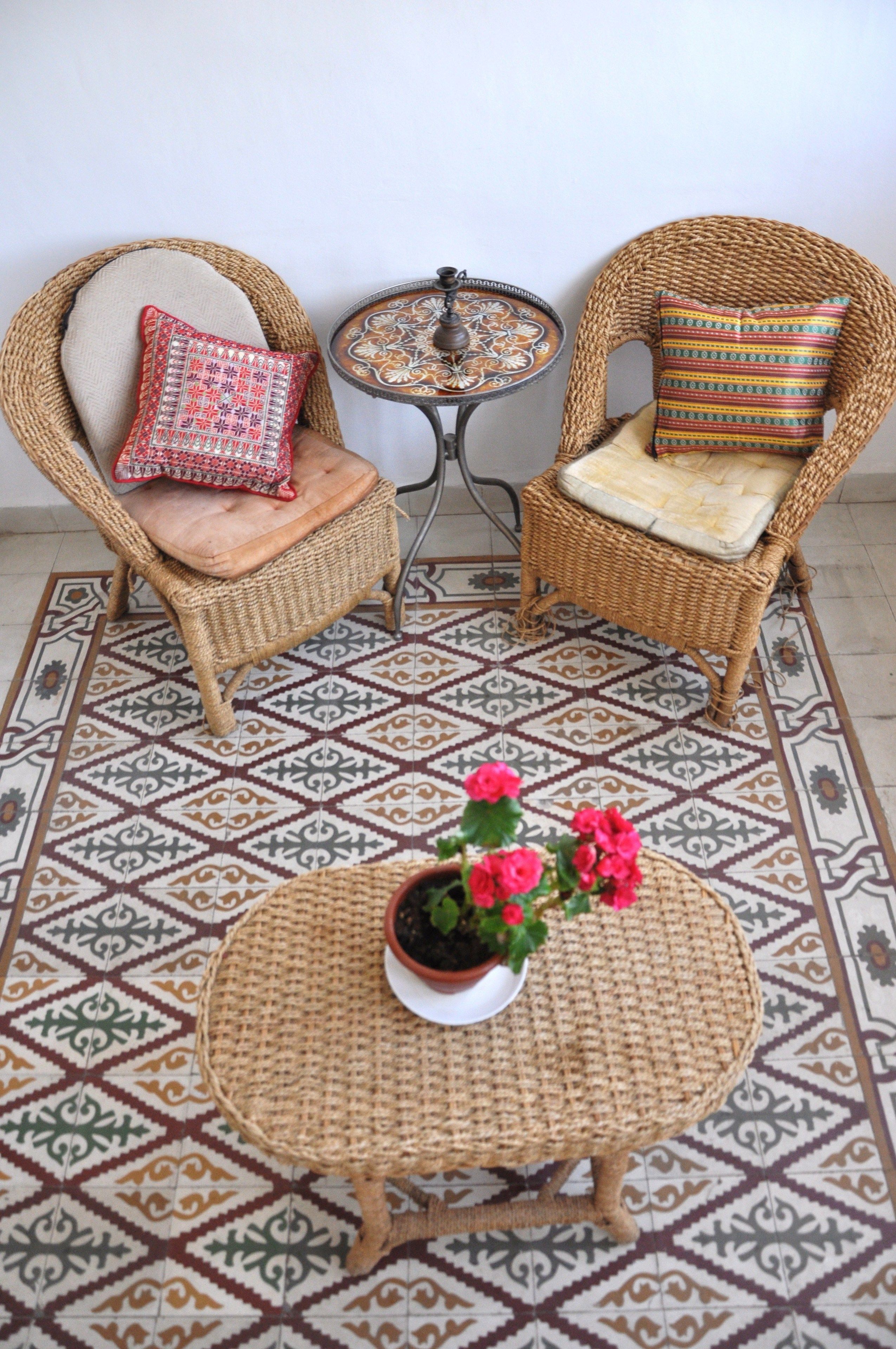 Quarto com duas cadeiras de vime, cada uma com almofada, uma pequena mesa redonda com uma planta em vaso e piso estampado.