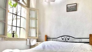 A neatly made bed with white linens, a window with a view of greenery, and a simple wall with a framed picture. - Al-Mutran Guest House (Nazareth)