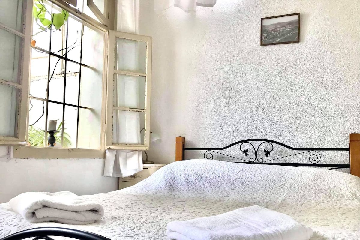 A neatly made bed with white linens, a window with a view of greenery, and a simple wall with a framed picture.