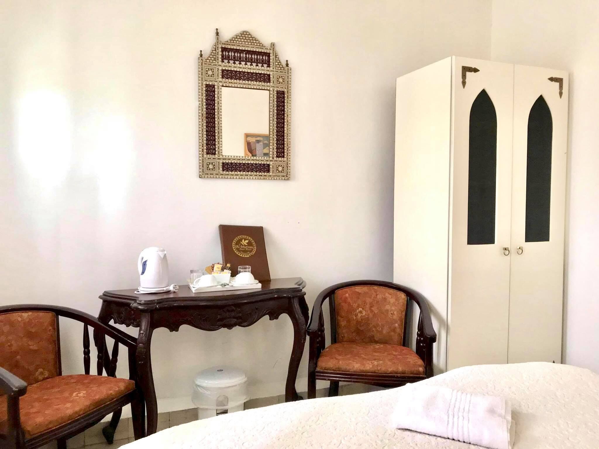 Una habitación con una cama, dos sillas, una mesa con una tetera, un espejo decorativo y un armario de dos puertas.