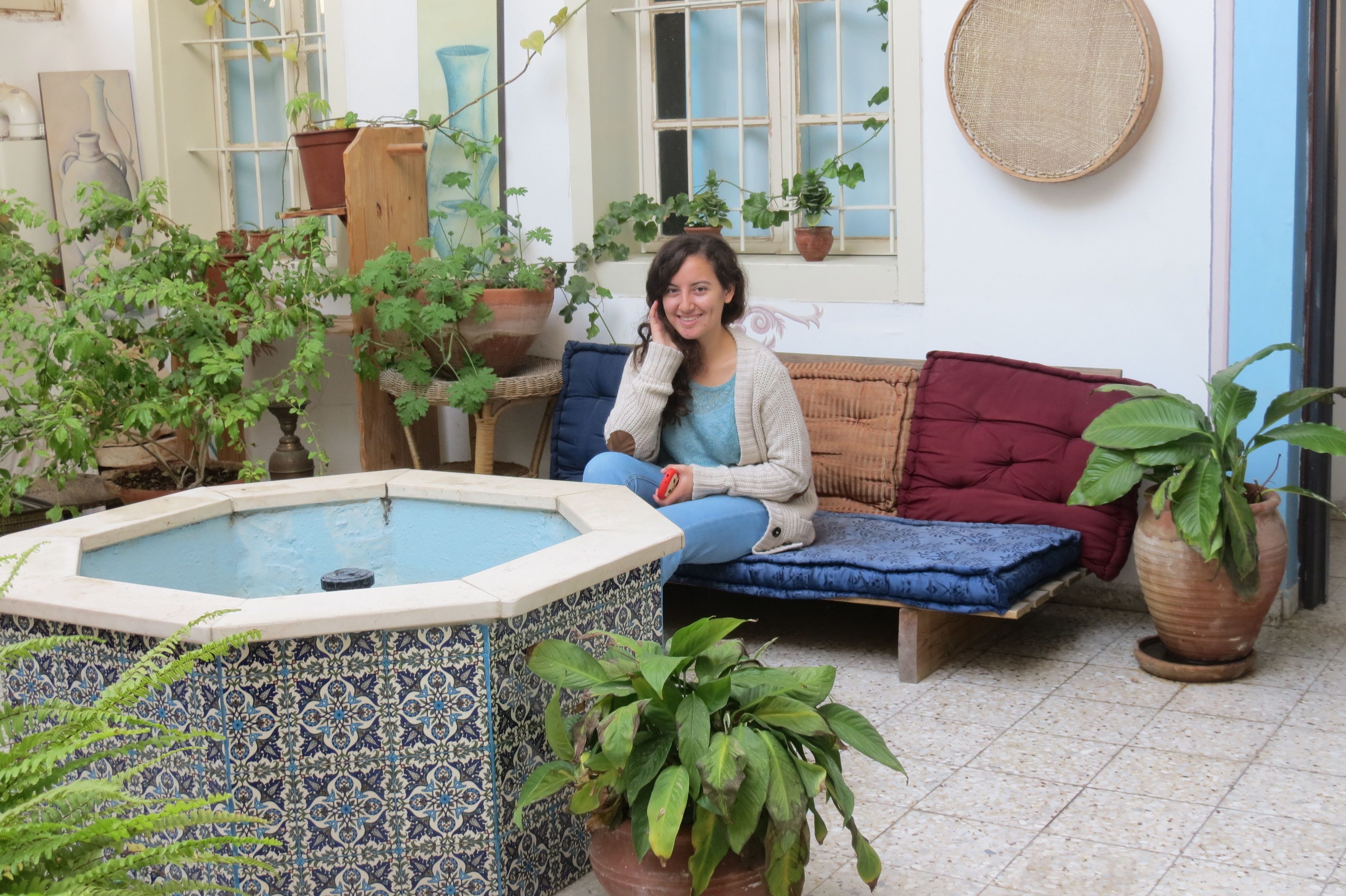 Eine Person sitzt auf einem blau-roten Sofa in einem Raum mit Fliesenboden, umgeben von Topfpflanzen und einer Zierbrunnen.