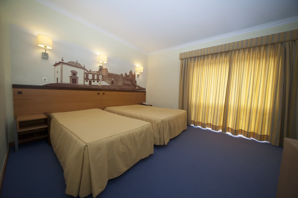 A hotel room with two single beds, a wooden headboard, a mural of a historic building, and a large window with curtains.