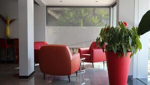 A modern lobby with red chairs, a large potted plant, and a glass wall with a view of greenery. - Hotel Feira Pedra Bela (Santa Maria da Feira)