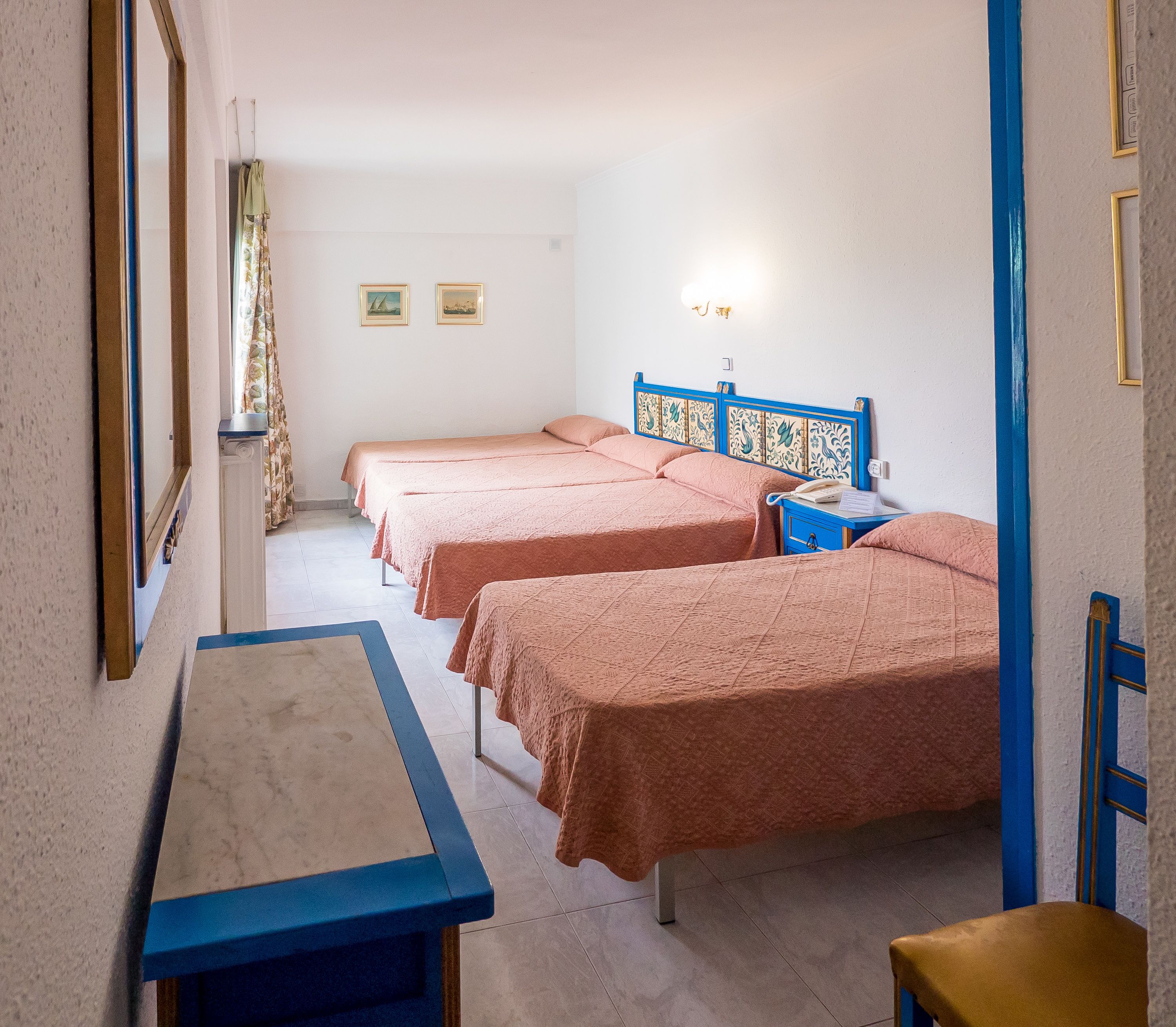 Une chambre avec trois lits, un bureau et une chaise. Deux tableaux encadrés sont accrochés au mur.