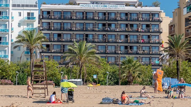 Ein Hotel mit mehreren Balkonen, ein Strand im Vordergrund und Palmen.
